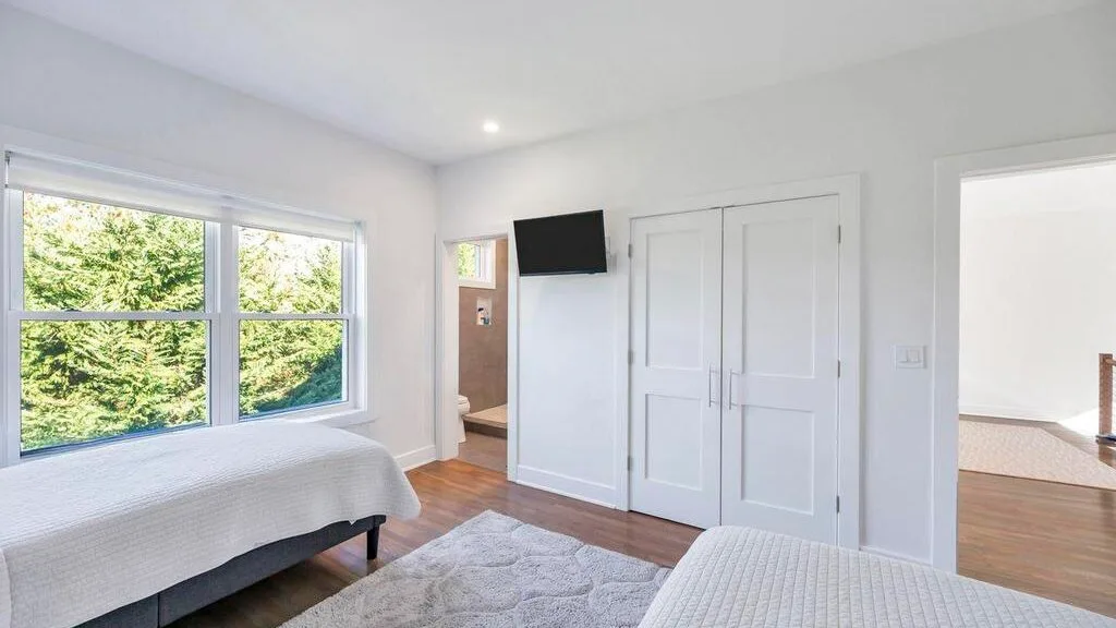 Un dormitorio luminoso con una cama junto a grandes ventanas y un televisor en la pared.