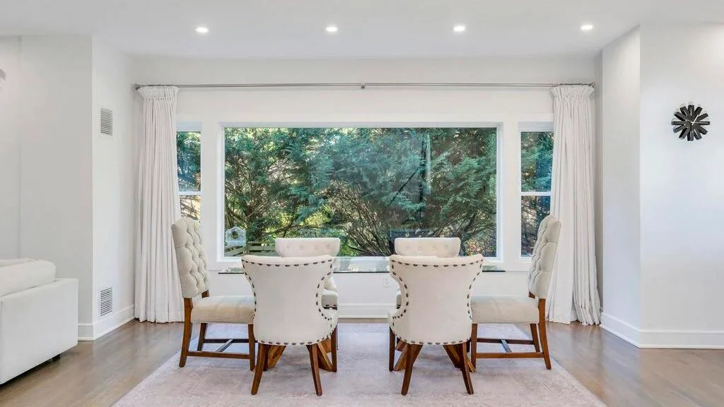 Un comedor luminoso con una mesa y asientos junto a grandes ventanas con vistas a la vegetación.