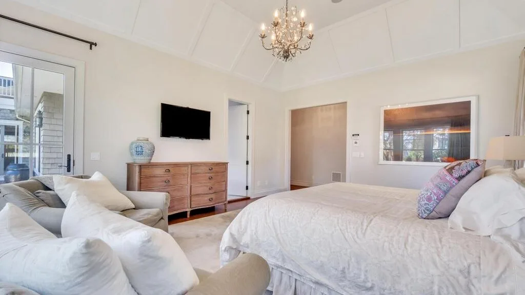 A bright bedroom with a large bed, a dresser, and a chandelier.