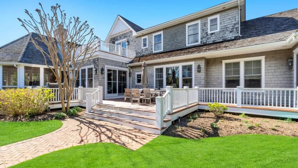 A large house opens onto a raised deck with outdoor seating and a green lawn.