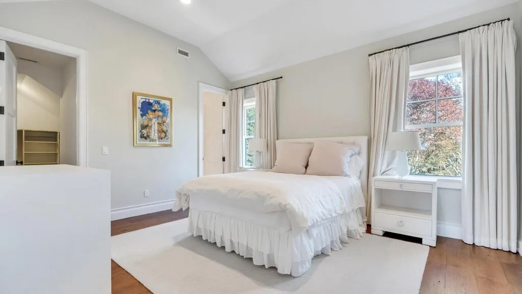 A bright bedroom with a white bed beside a large curtained window.