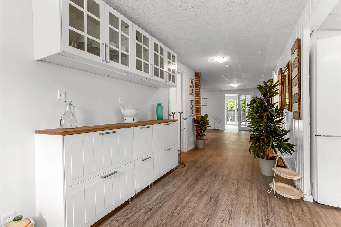 Un pasillo luminoso con armarios blancos, suelo de madera y plantas en macetas.