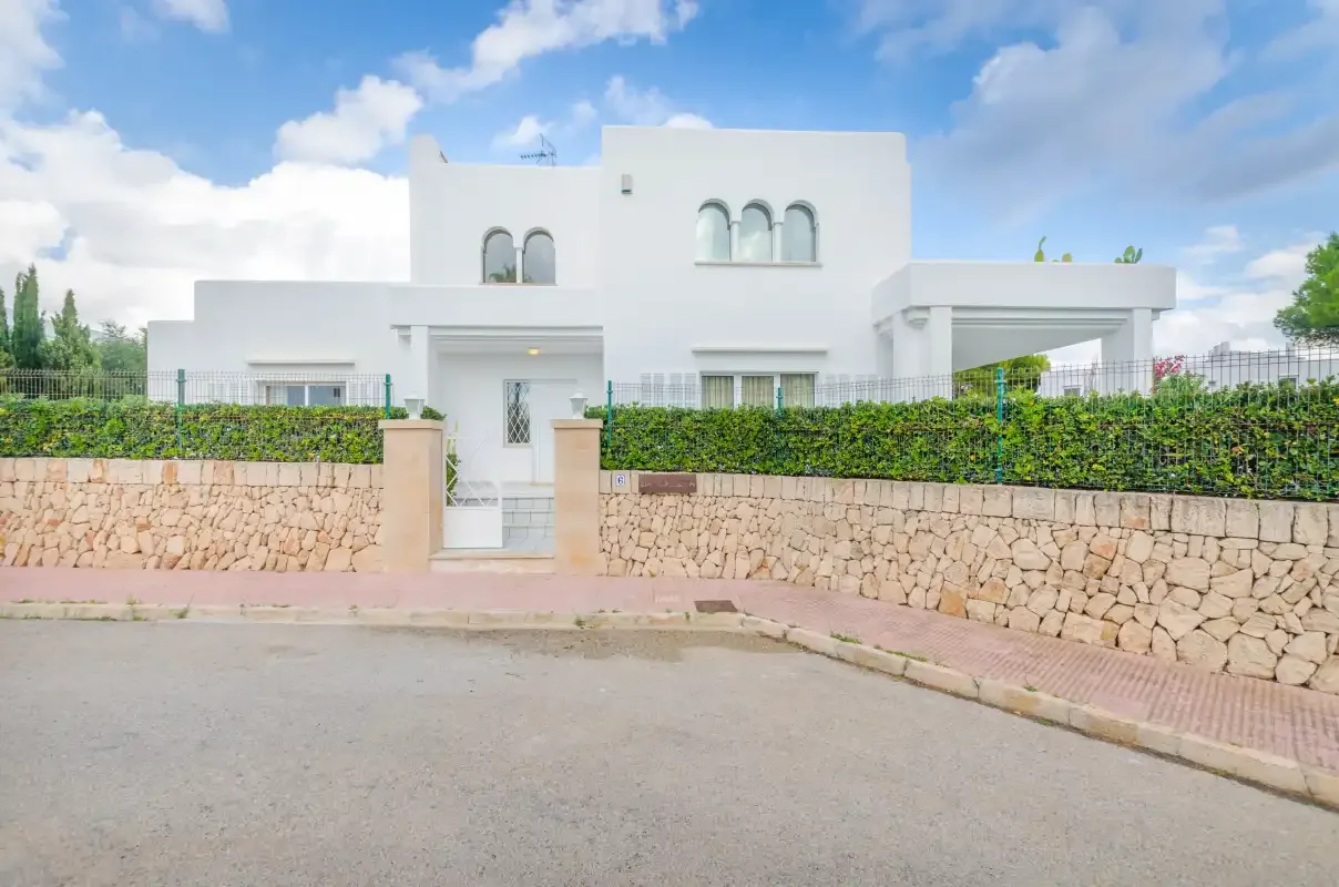 A white modern house stands behind a low stone wall and hedge.