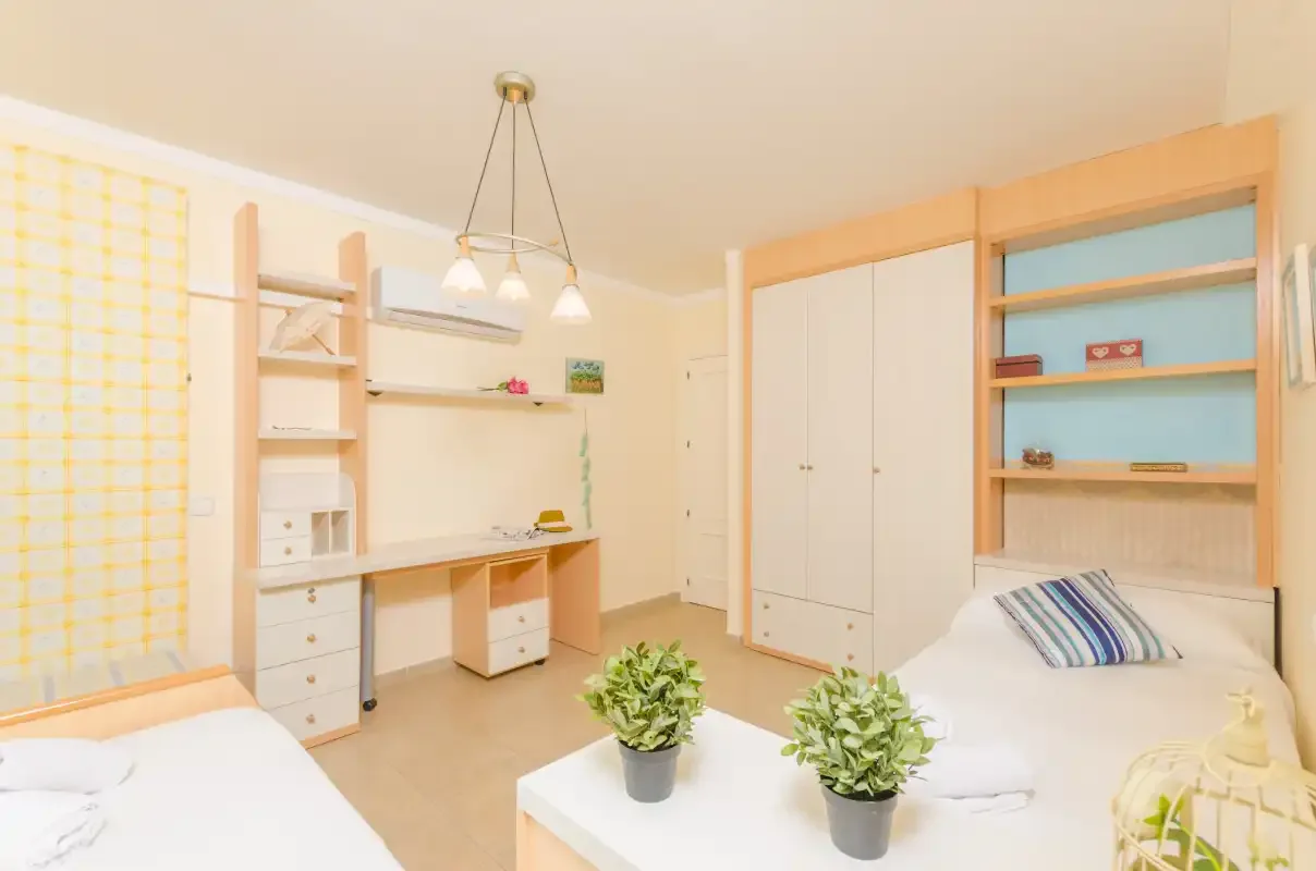 A bright bedroom with two beds, a desk, and built-in shelving.