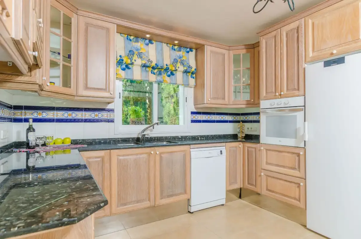 A kitchen with light wood cabinetry, dark countertops, and a window above the sink.