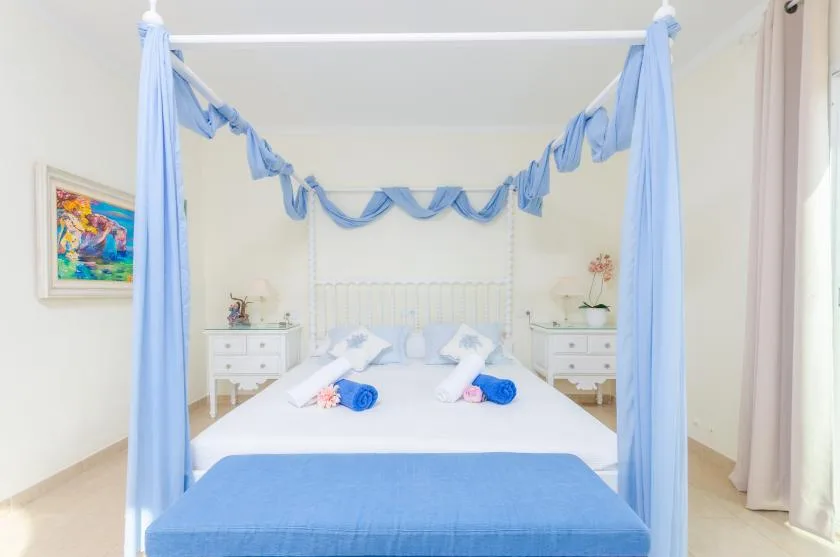 A bright bedroom features a white bed with blue canopy drapes and a blue bench.