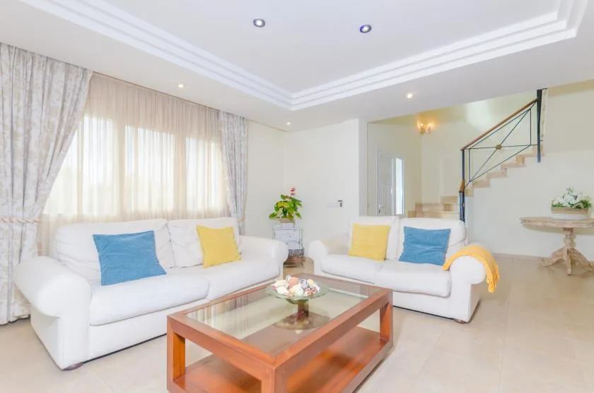 A bright living room with white seating, colorful cushions, and a glass-topped coffee table.