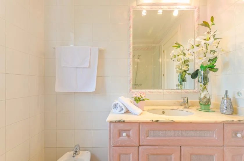A bright bathroom with a sink, vanity, and flowers beside the mirror.