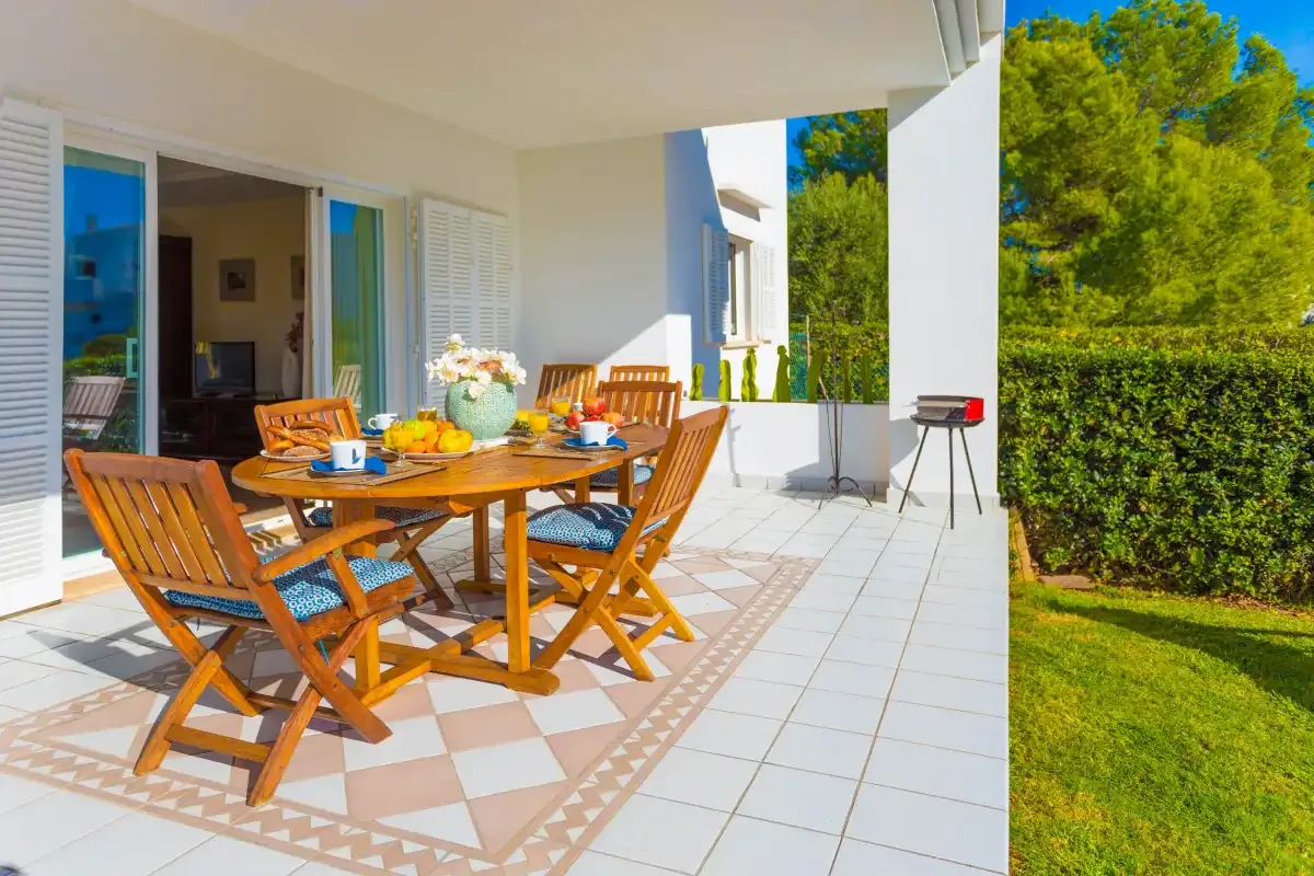 A covered outdoor dining area has a wooden table and chairs beside a green garden.