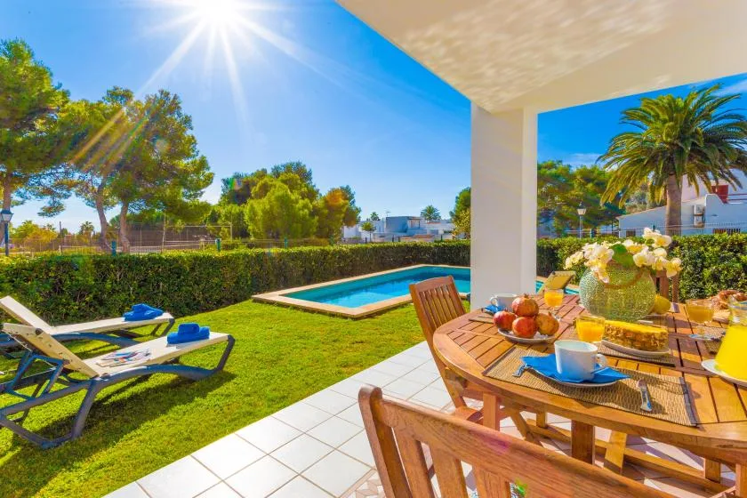 A sunny patio dining area overlooks a lawn, swimming pool, and surrounding trees.