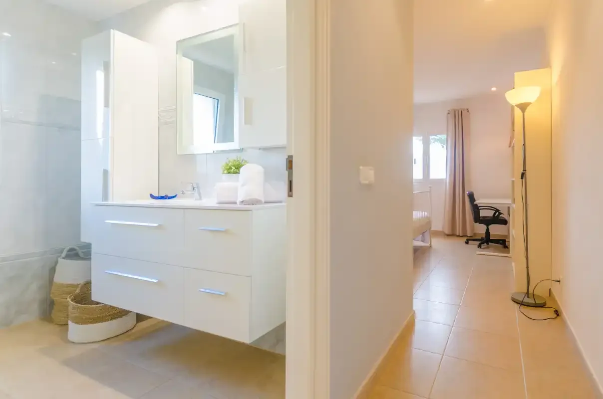 A bathroom vanity opens onto a hallway leading to a bedroom with a desk.