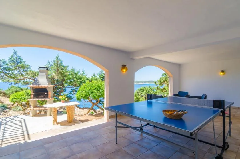 A covered patio with a ping-pong table overlooks a garden and the water.