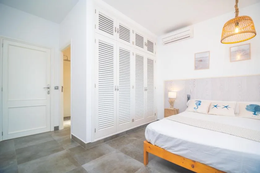 A bright bedroom with a bed, white louvered closet doors, and a woven pendant light.