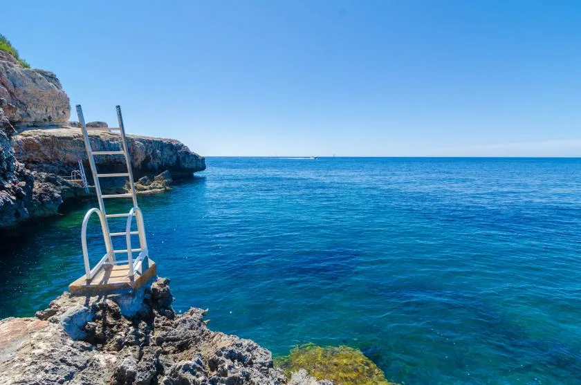 A rocky shoreline meets clear blue sea, with a metal ladder beside the water.