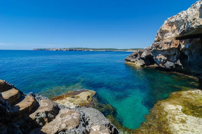 A rocky coastline borders clear blue water under a bright sky.