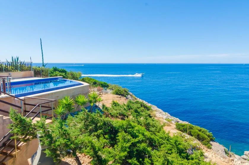 A swimming pool and surrounding greenery overlook the ocean beneath a clear sky.