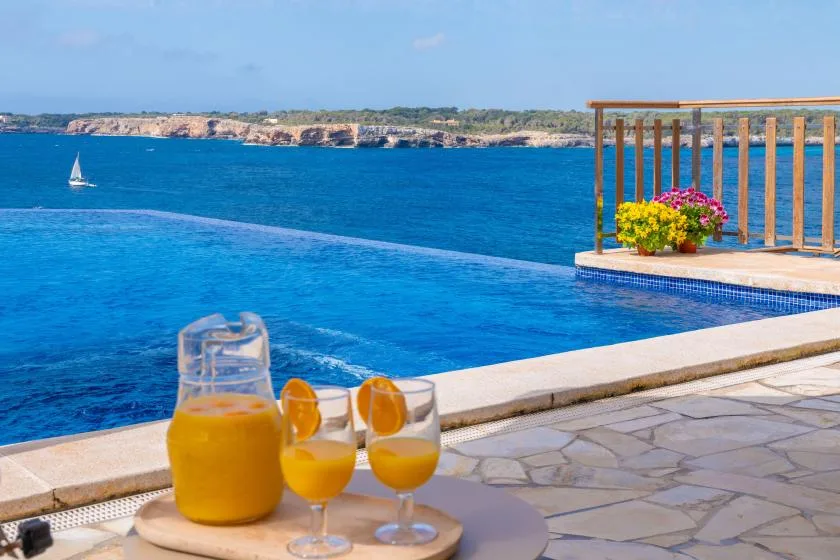 A pool overlooks the blue sea, with drinks on a table beside the water.