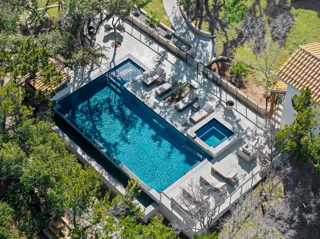 Aerial view of a rectangular outdoor pool with adjacent hot tub, surrounded by lounge chairs, trees, and a modern patio area.