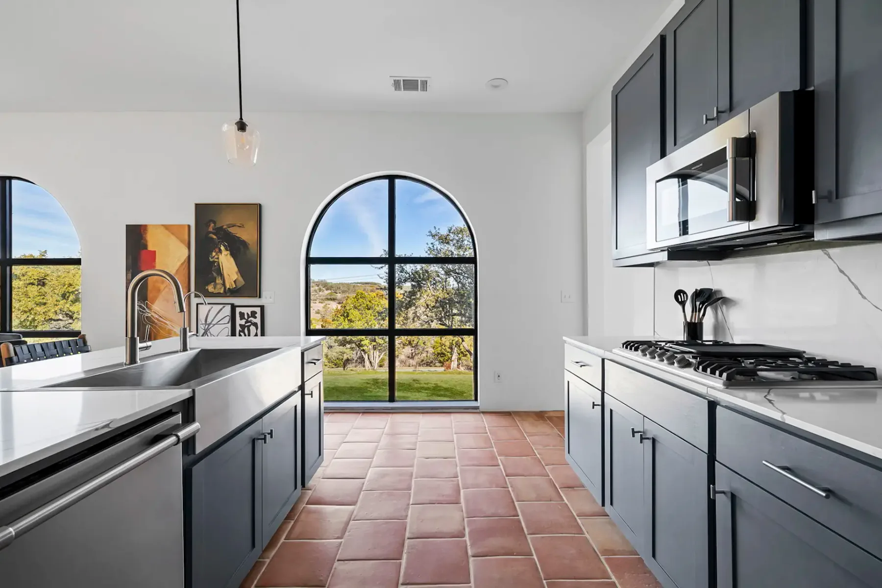 Modern kitchen with arched windows, blue cabinets, stainless steel appliances, white countertops, and terracotta tile flooring.