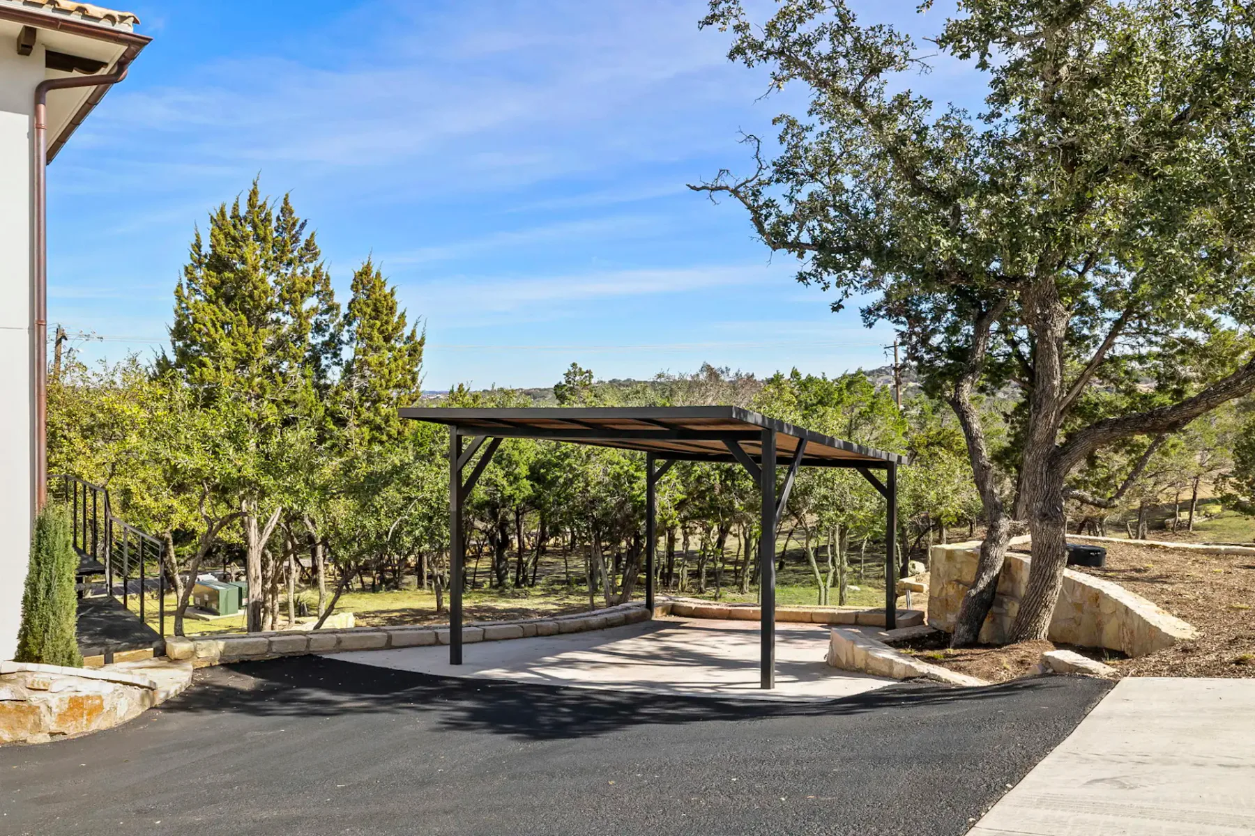 A metal-roofed carport stands beside a house, with trees and paved ground nearby.