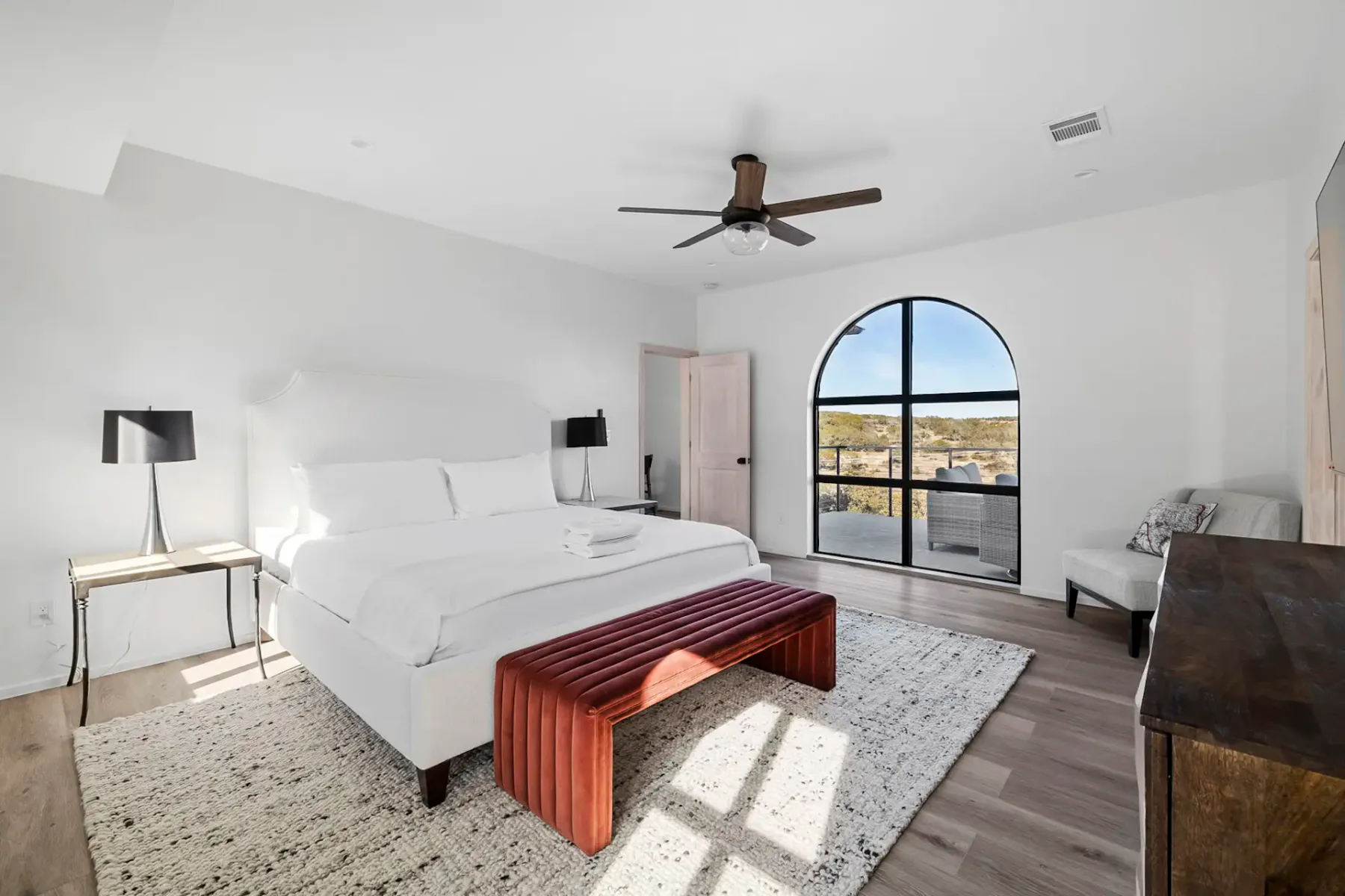 Minimalist bedroom with a white bed, red bench, arched window, ceiling fan, and modern lamps on side tables. Light wooden floor and rug.