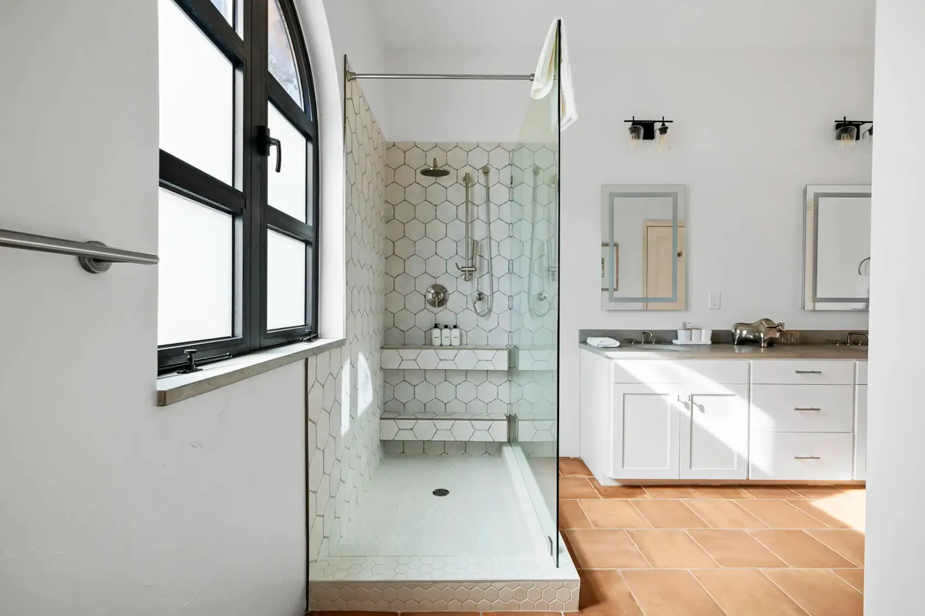 A bright bathroom with a glass shower, white vanity, and terracotta-colored floor tiles.