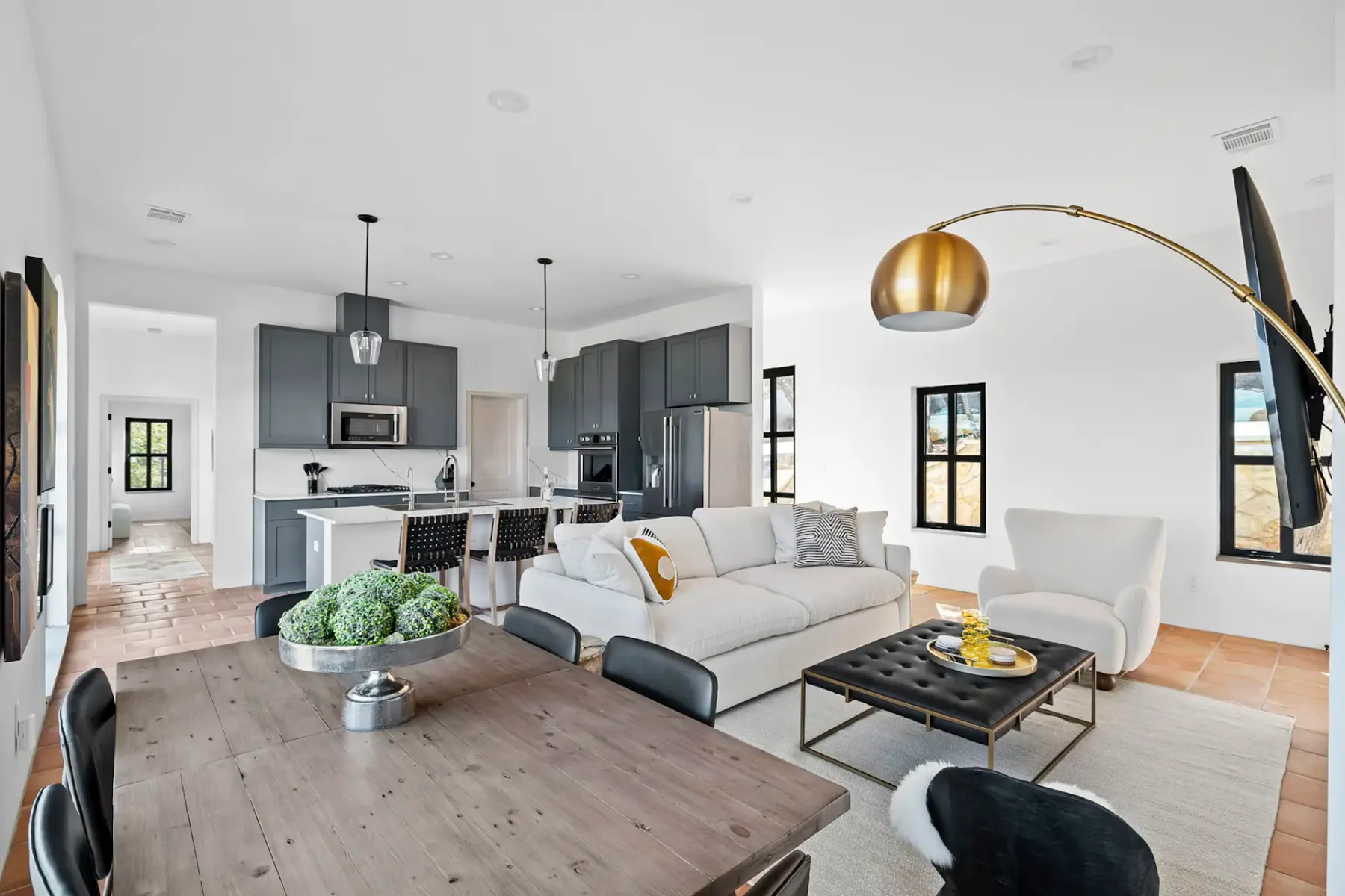 Modern open living room and kitchen with gray cabinets, white walls, and terracotta tile floor. Featuring a dining table, sofa, and armchair.