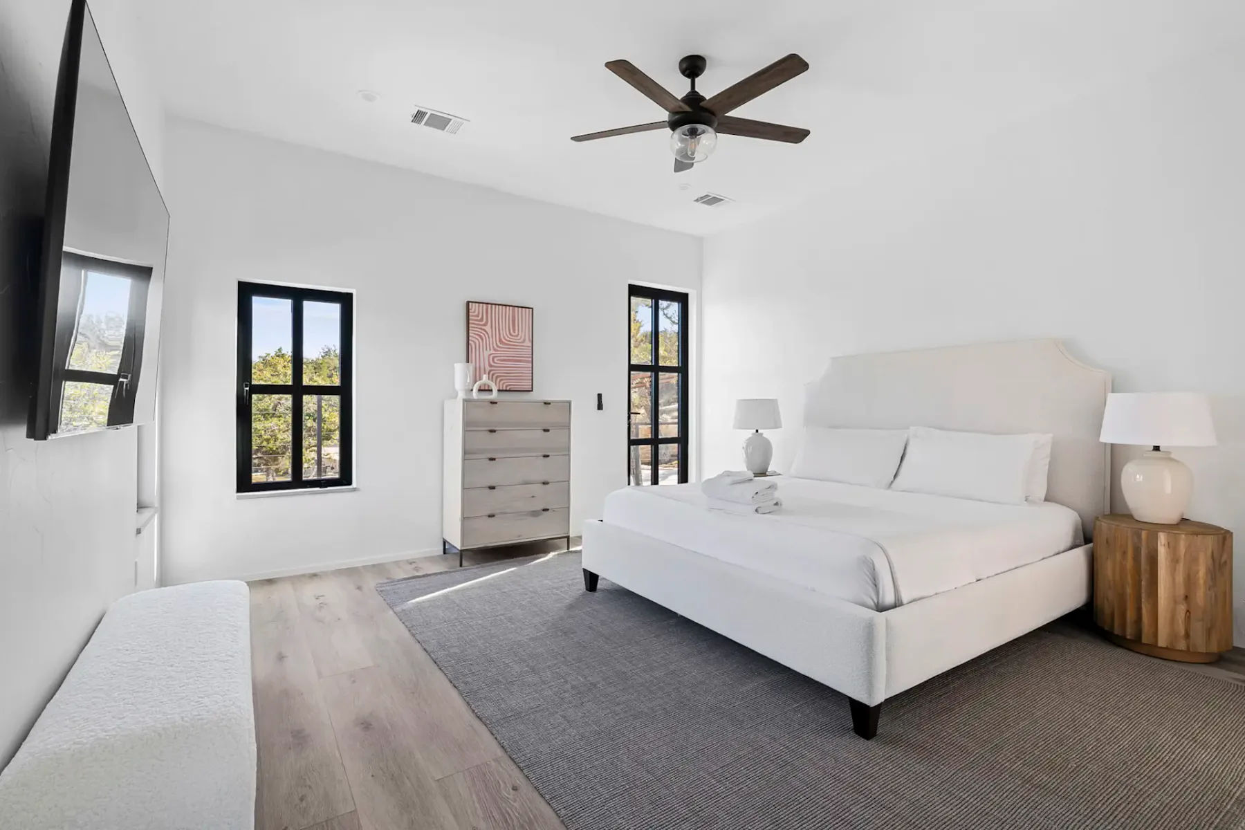 Minimalist bedroom with a large bed, white walls, wooden furniture, a ceiling fan, and sunlight streaming through two windows.