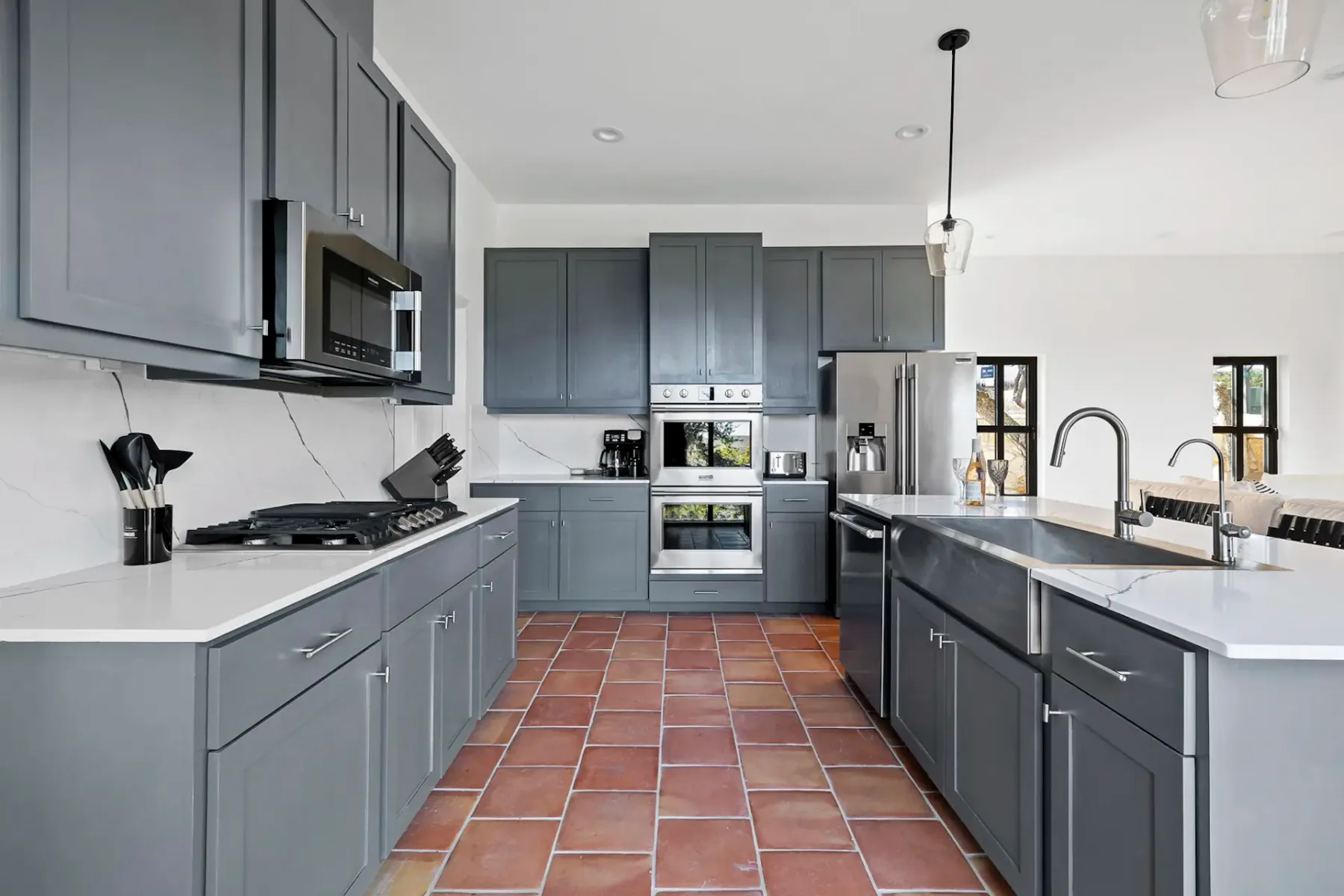 Modern kitchen with gray cabinets, stainless steel appliances, a double sink, and terracotta tile floor. Bright lighting and sleek design.