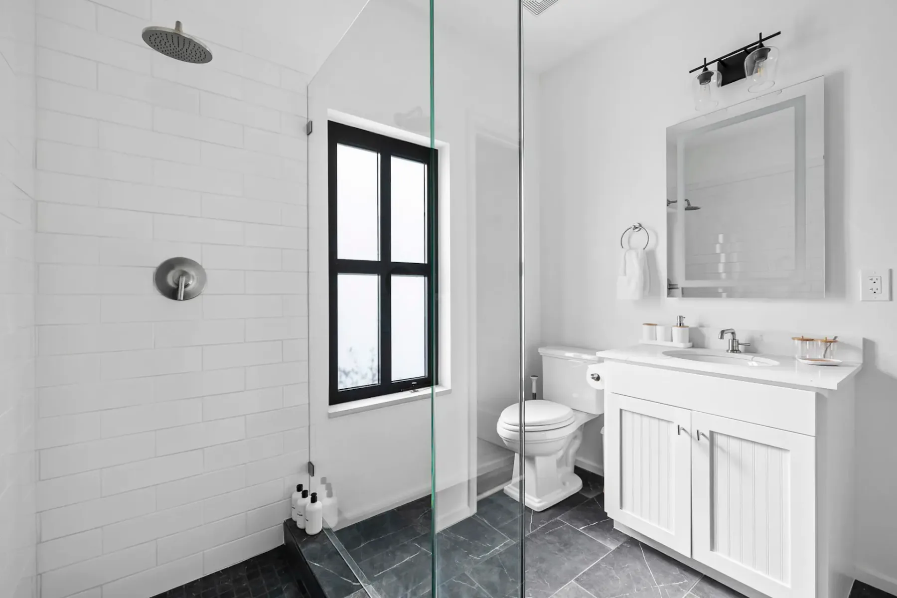 A modern bathroom with a glass shower, white vanity, toilet, and black-framed window. White walls and dark tile flooring.