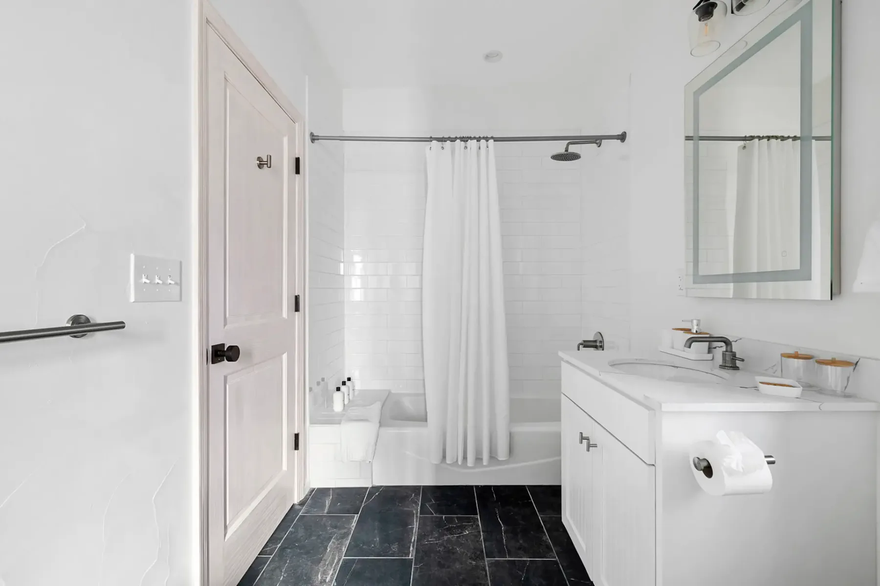 A clean, white bathroom with a bathtub, shower curtain, sink, mirror, and black tiled floor.