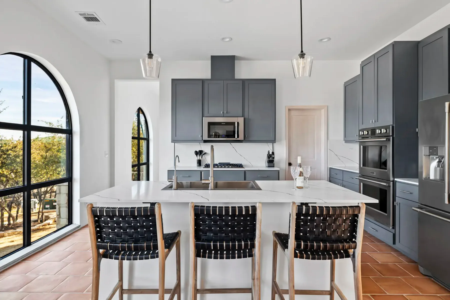Modern kitchen with gray cabinets, a white island, and black woven chairs. Arched windows with a garden view enhance natural light.