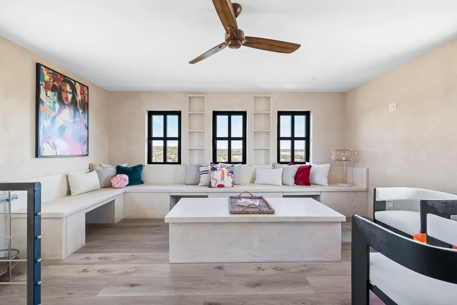 Bright, minimalist living room with large windows, beige walls, wooden floor, ceiling fan, bench seating, cushions, and colorful artwork.