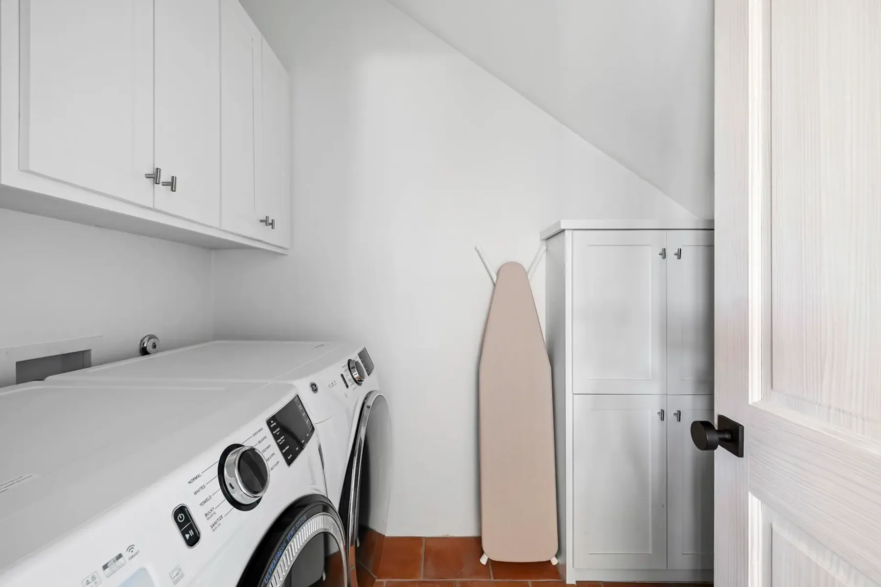 Laundry room with white washer and dryer, wall cabinets, a white storage cupboard, and an ironing board. Brown tiled floor.