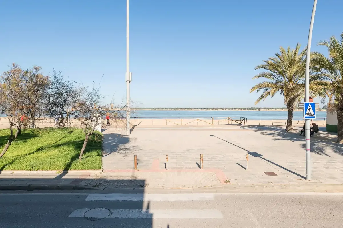 A seaside promenade opens onto a sandy beach, with palms and a pedestrian crossing sign.