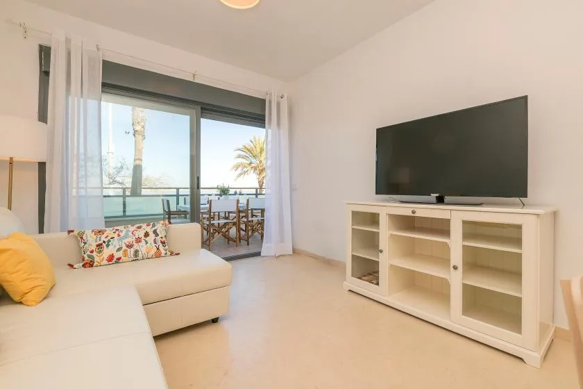A bright living room has white seating, a television, and sliding glass doors to a balcony.
