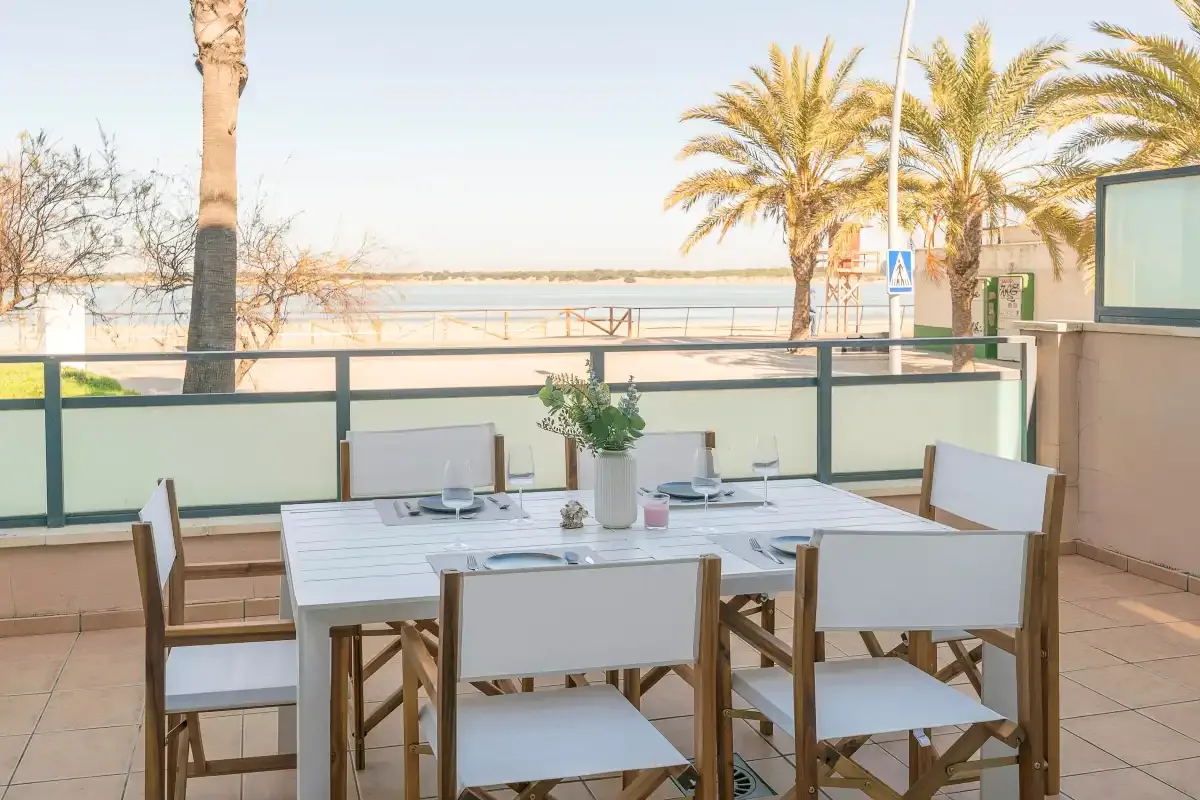 A furnished outdoor dining terrace overlooks a waterfront promenade and palm trees.