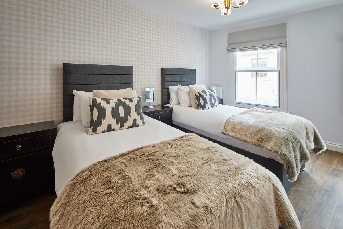 A bright bedroom with two beds, patterned pillows, and light-colored throws.