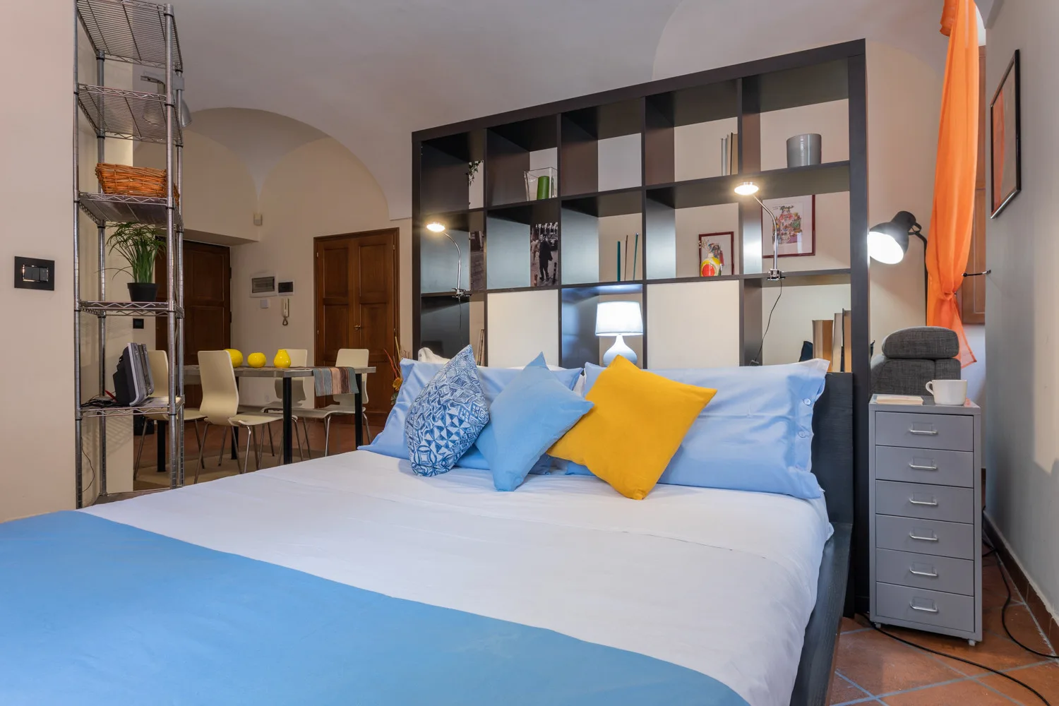 Un dormitorio con una cama vestida de blanco y azul, frente a una estantería abierta.