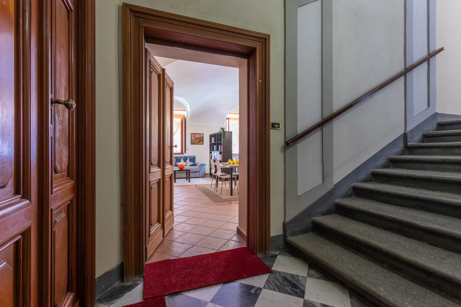 Un pasillo interior con una escalera junto a una puerta abierta hacia una habitación amueblada.