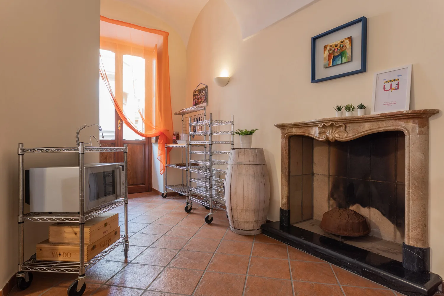 Una habitación con suelo de baldosas, una chimenea grande, estanterías metálicas y una ventana con cortinas naranjas.