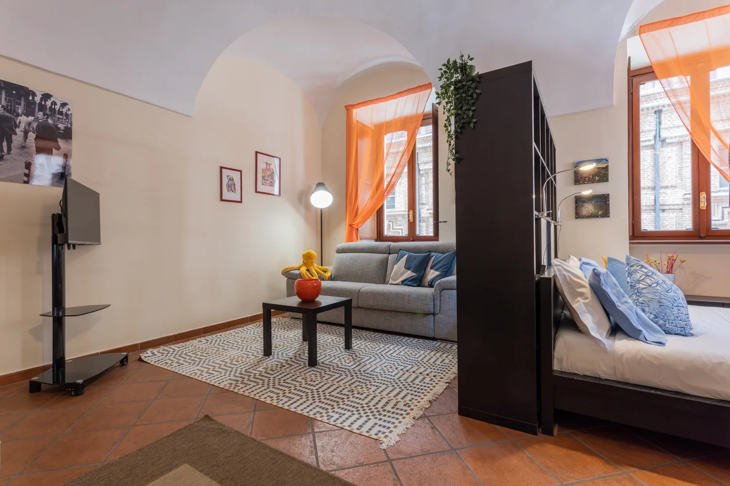A living area with a gray sofa, coffee table, and orange curtains beside a bed.