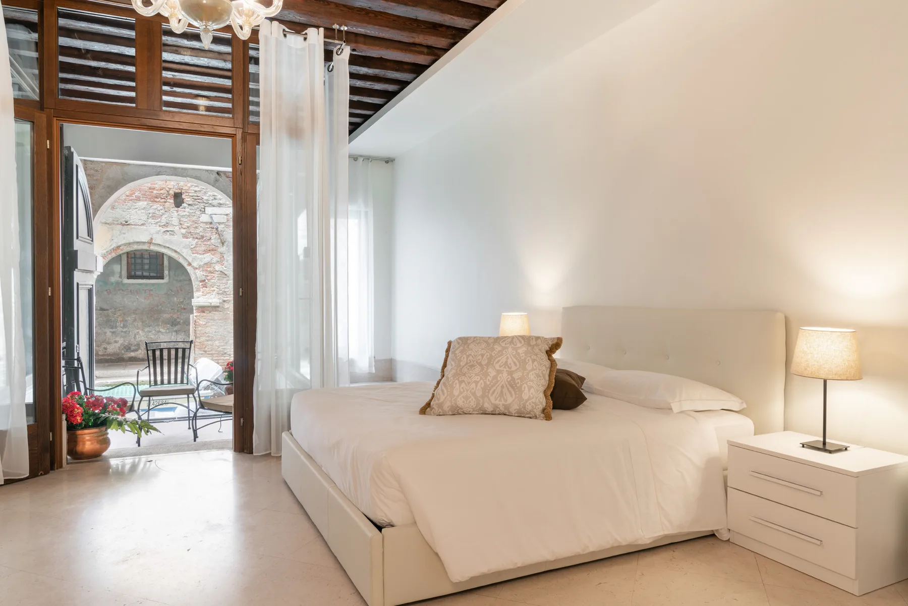 Un dormitorio luminoso con una cama grande junto a ventanas altas y una puerta abierta al balcón.