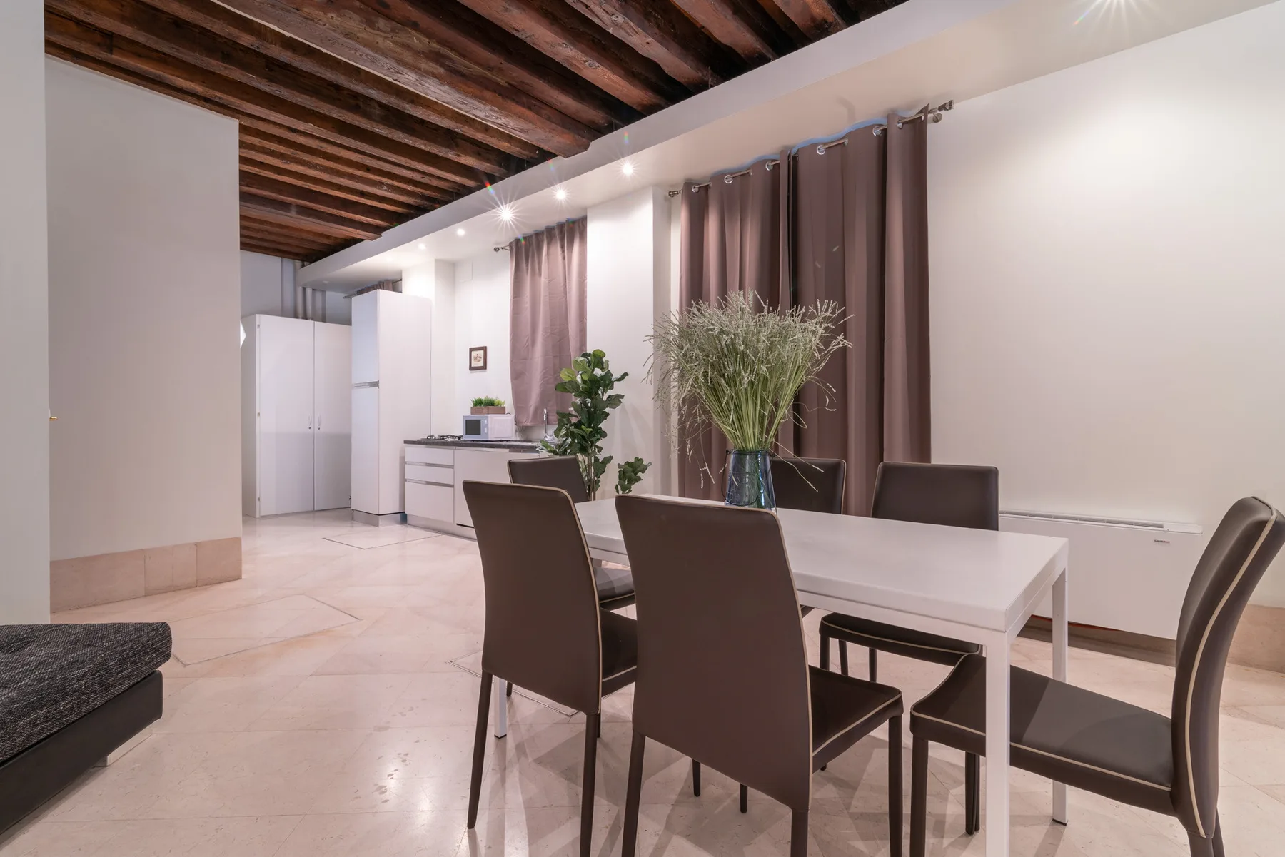 Un comedor con una mesa blanca y asientos tapizados oscuros bajo vigas de madera expuestas.