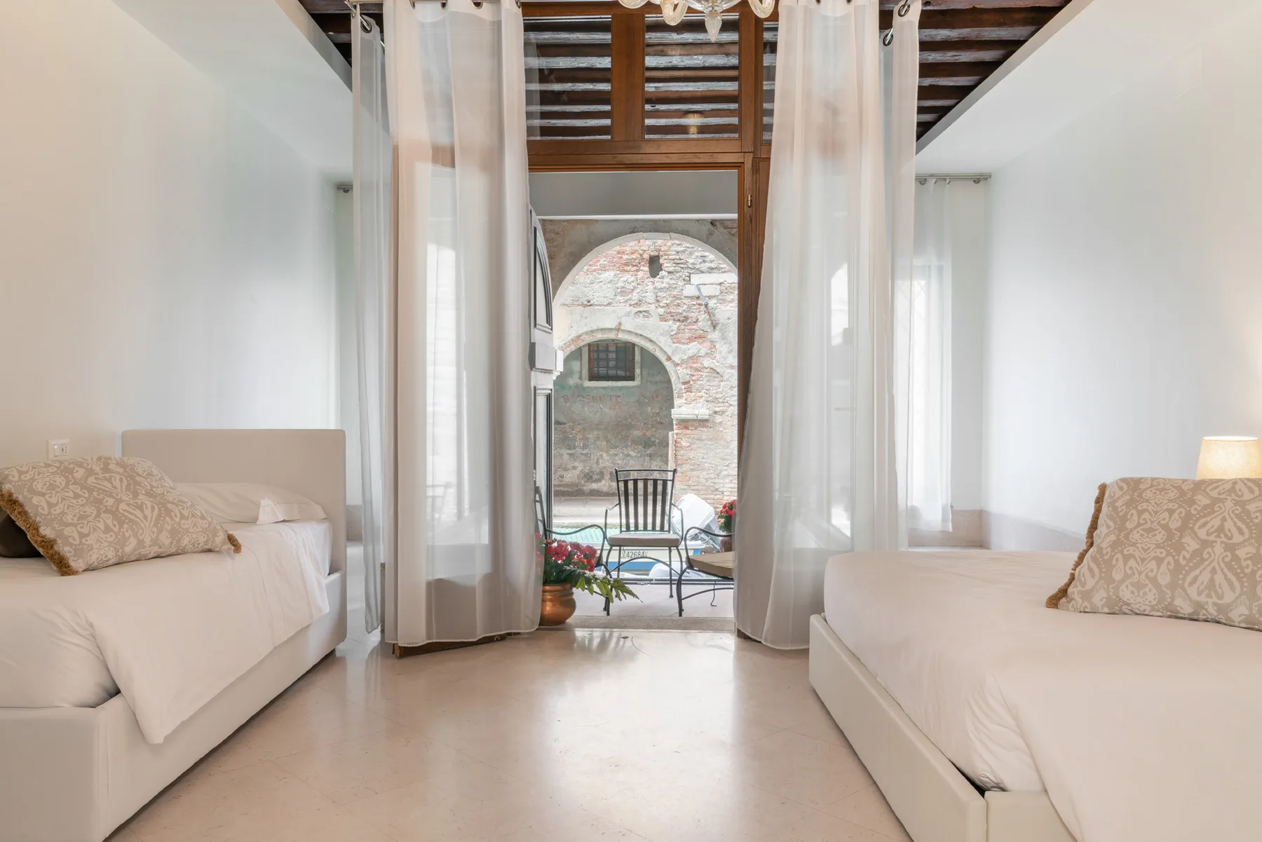 Un dormitorio luminoso con dos camas y cortinas translúcidas que enmarcan una puerta arqueada.