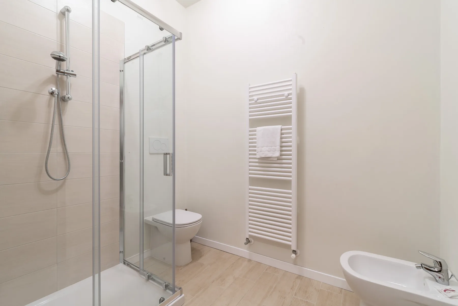 Un baño luminoso con ducha de cristal, inodoro y radiador toallero de pared.
