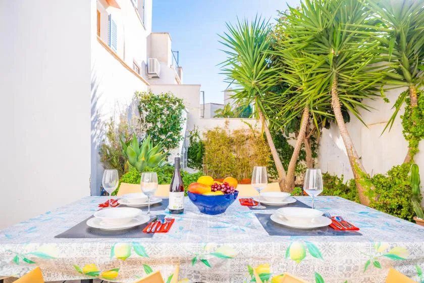 An outdoor dining table is set beneath leafy trees beside a white building.