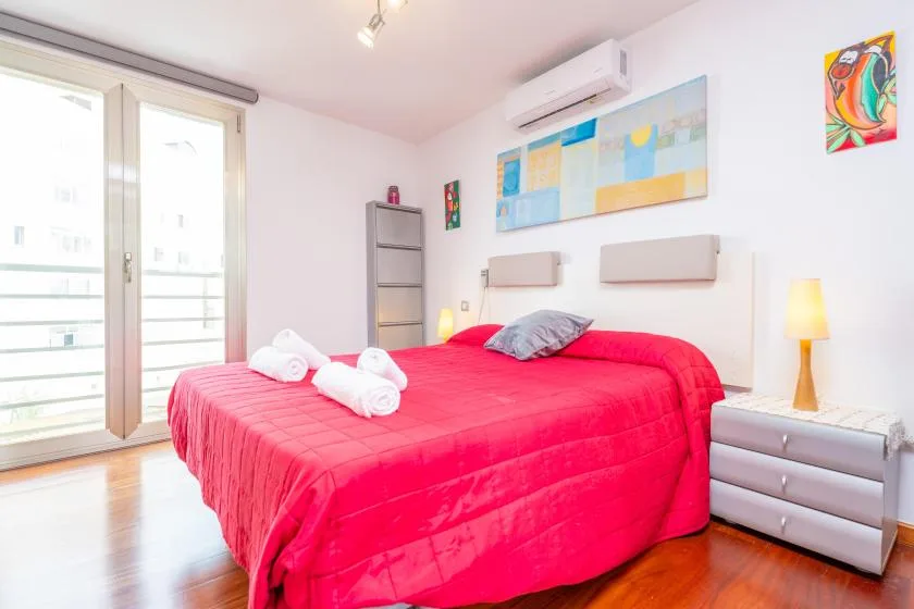 A bright bedroom features a large bed with vivid red bedding and a balcony door.