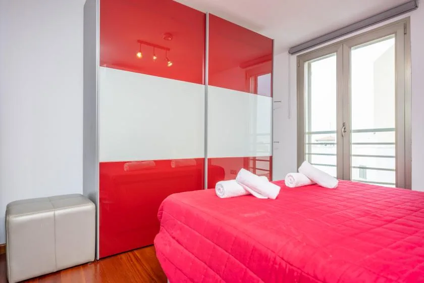 A bedroom with a bright red bed beside a large window.