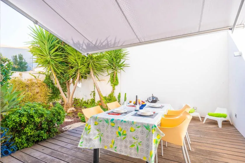 A covered outdoor dining area has a table beside leafy plants and wooden decking.